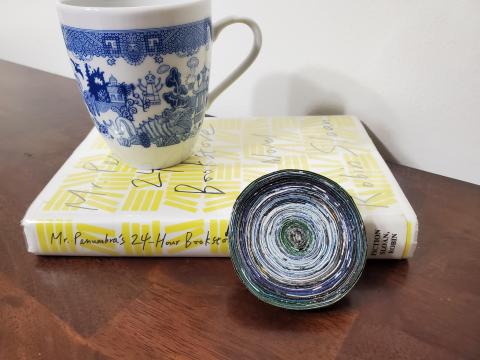 A blue and white coffee mug sits upon a book with a coiled magazine coaster leaning against the book's spine.