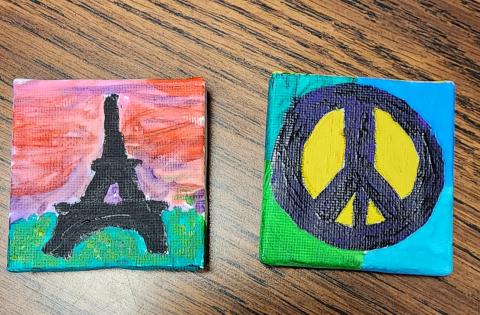 Two 2-inch canvases are on display. One painting is of the Eiffel Tower with a pink sky and green grass. The second painting is purple peace sign with yellow in-between the stems and blue and green background. 