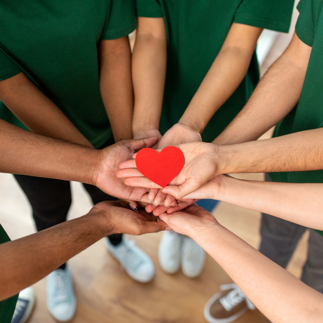 Many hands extended to join in a circle holding a paper heart cut out.